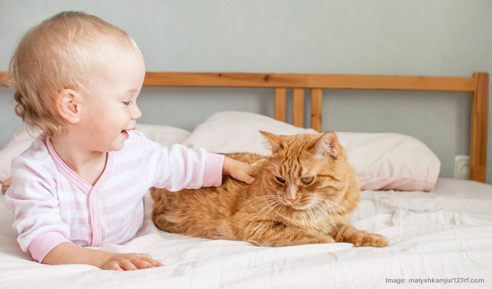 Baby and ginger cat laid next to each other on a bed, the baby is stroking the cat.