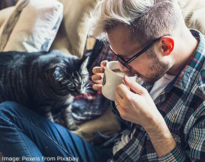 Man on sofa drinking from a cup with a cat standing next to him, photo by pexels on pixabay.
