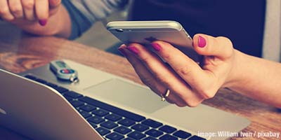 Person with red fingernails sitting and looking at a phone and computer.